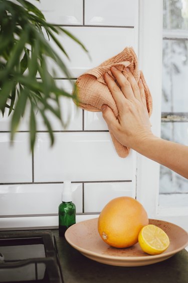 Person using a pink washcloth to clean a white subway tile backsplash, and a counter with a bottle and a plate of oranges
